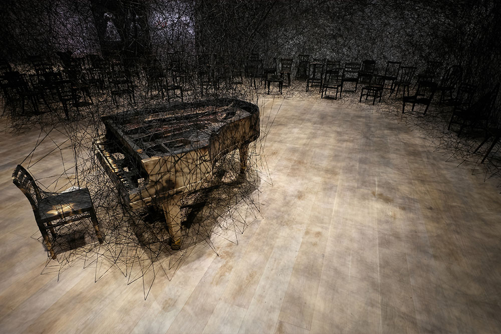 Burnt piano and chair stand in a room. Behind them are an array of chairs connected to each other and the stairs via black wool