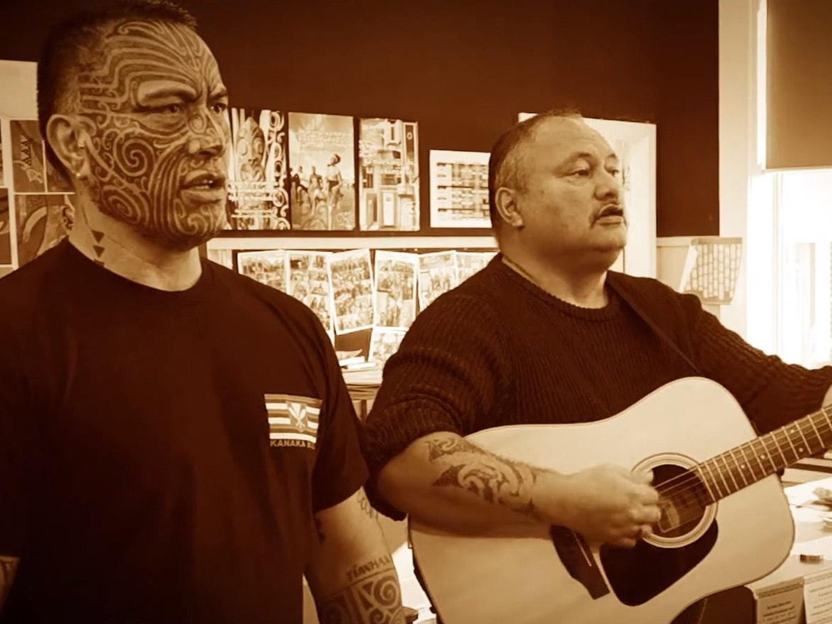 Two men singing with a guitar, one man has a full-face tattoo