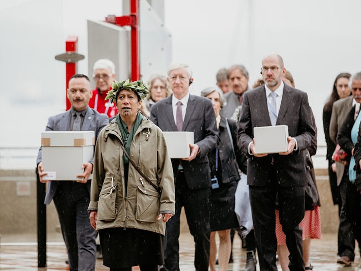 The delegation prepares to enter Te Papa’s marae. Te Papa’s Lisa Osborne gives the traditional karanga (call). Visible carrying three of the four toi moko are (left to right) Te Papa Repatriation Coordinator Te Arikirangi Mamaku, German Ambassador to New A group of people enter a building. Three of them carry boxes