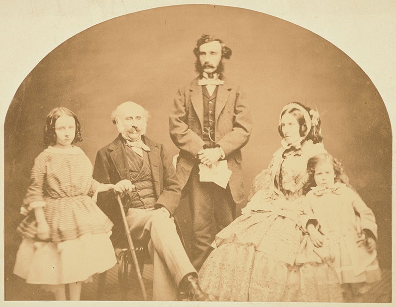 Family portrait. A man stands in the centre. He is flanked by an older man and an older woman, both sitting. They are flanked by two children