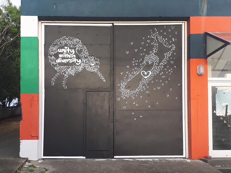 A black sign on a garage that has a white kiwi and a map of New Zealand drawn on it with the words "Unity within diversity"