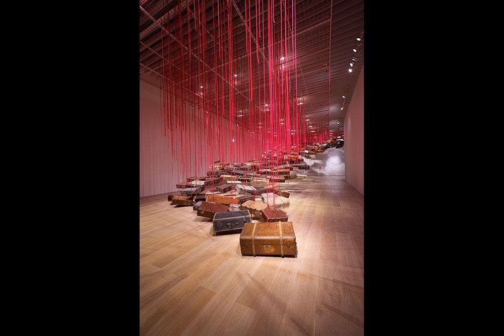 Large room with suitcases suspended from the ceiling by red thread forming a pathway to the roof