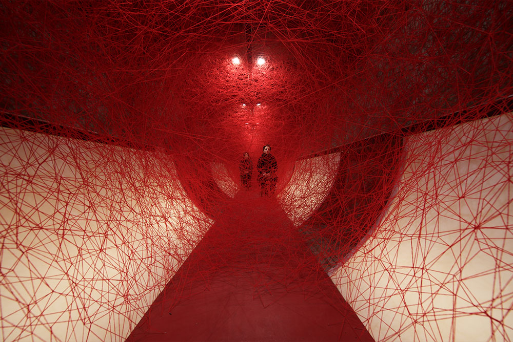 Room shaped like a tube containing multiple strands of red fibre woven together across it to resemble a giant web. Behind the web sit two people on chairs