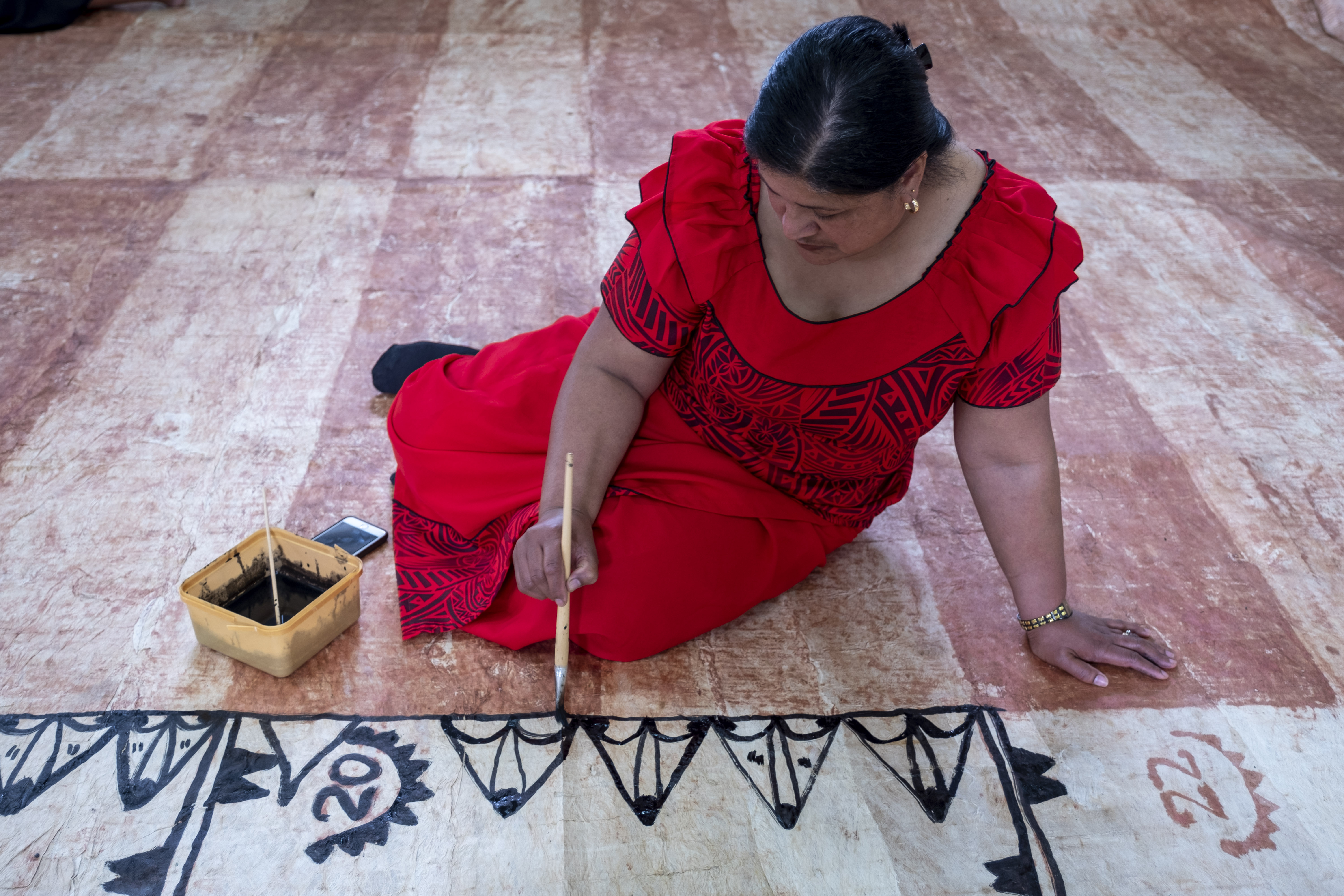 A woman in a red dress painting black ink on a tapa cloth on the floor