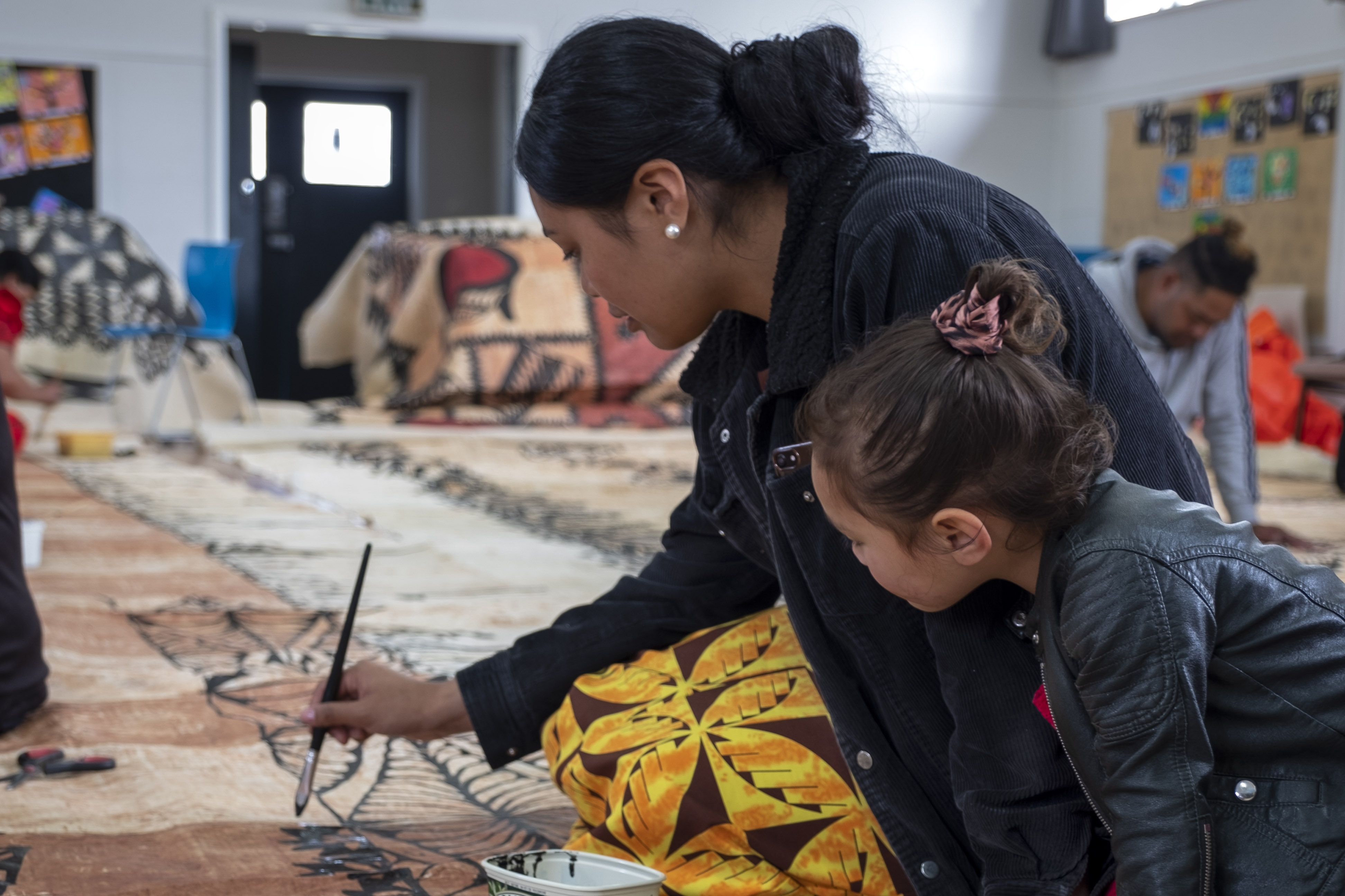 A woman painting black ink on a tapa cloth on the floor, she is being watched by a child.