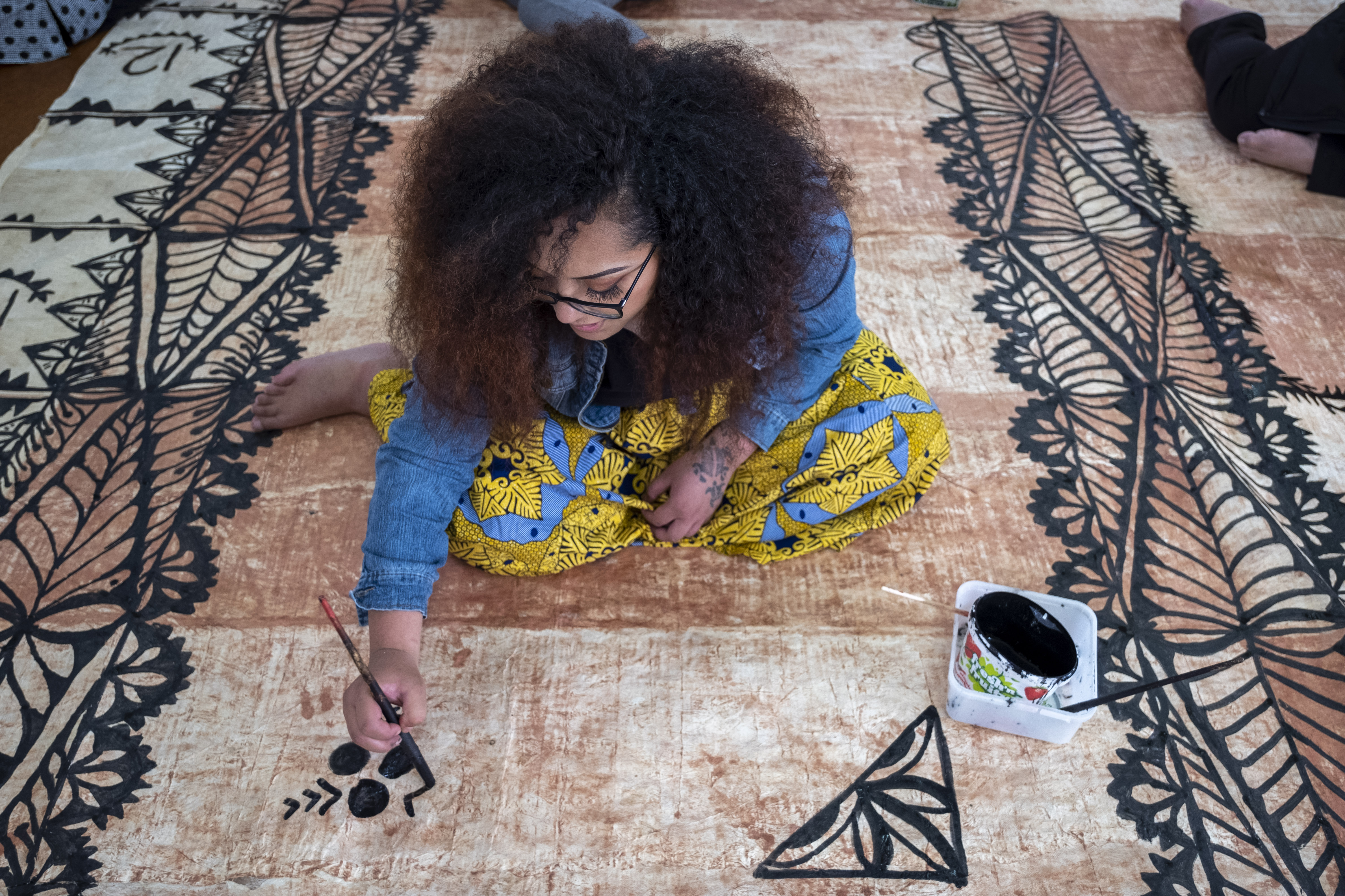 A woman painting black ink on a tapa cloth on the floor.