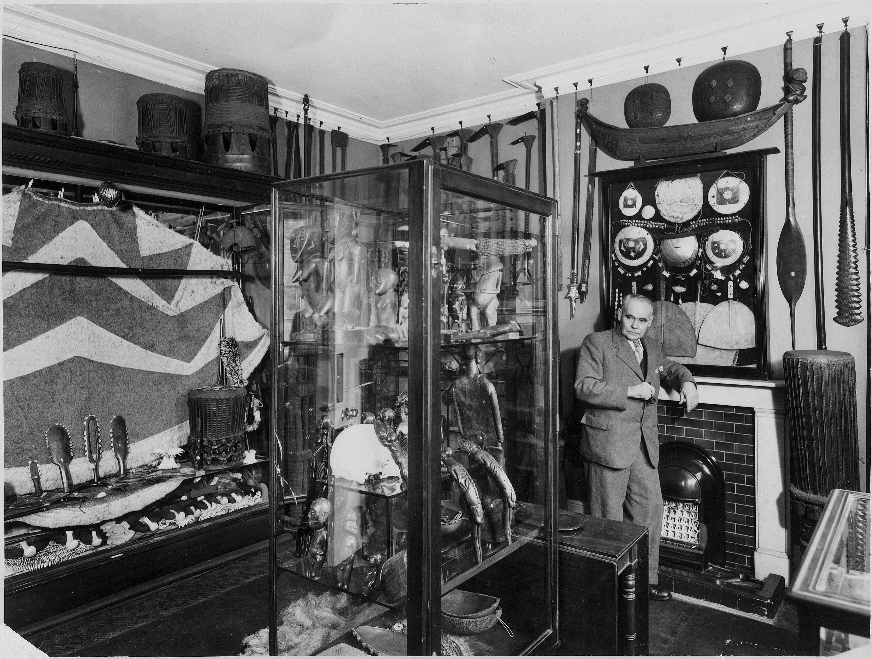 A black and white photo with an older man standing in the middle of a lot of traditional Pacific art pieces