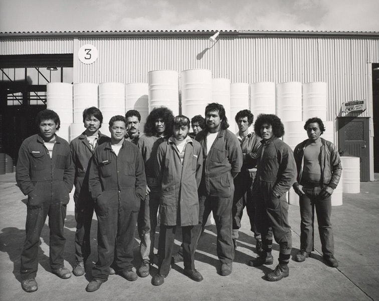 Black and white photo of 11 men in overalls standing in front of a factory