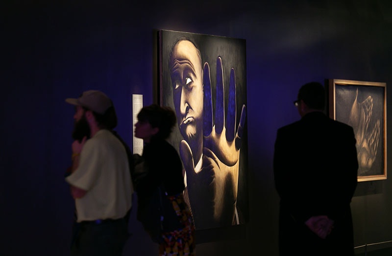 People look at a painting by Tony Fomison. The painting depicts a man holding his hand up to the camera as if to say No