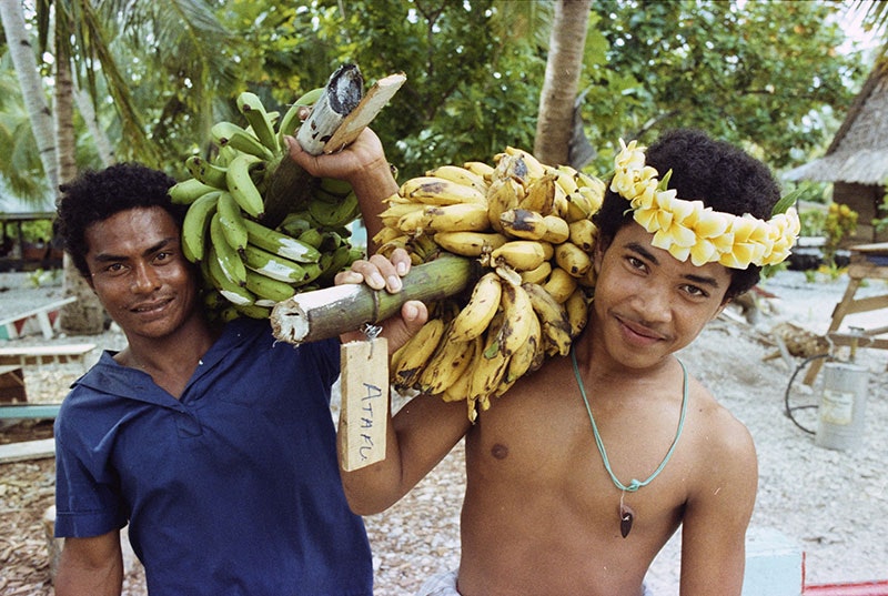 Two men holding bunches of bananas balanced on their shoulders pose for a photo