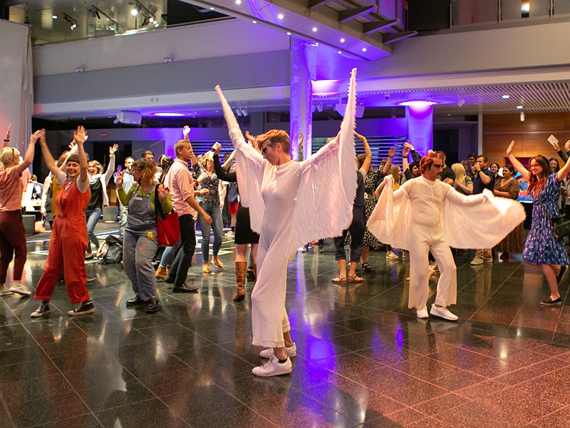 People dancing in a large space. Two of the people are in white outfits that stretch over long 'arms' much longer than their real arms.