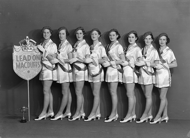 Black and white photo of women lined up beside a trophy in uniform