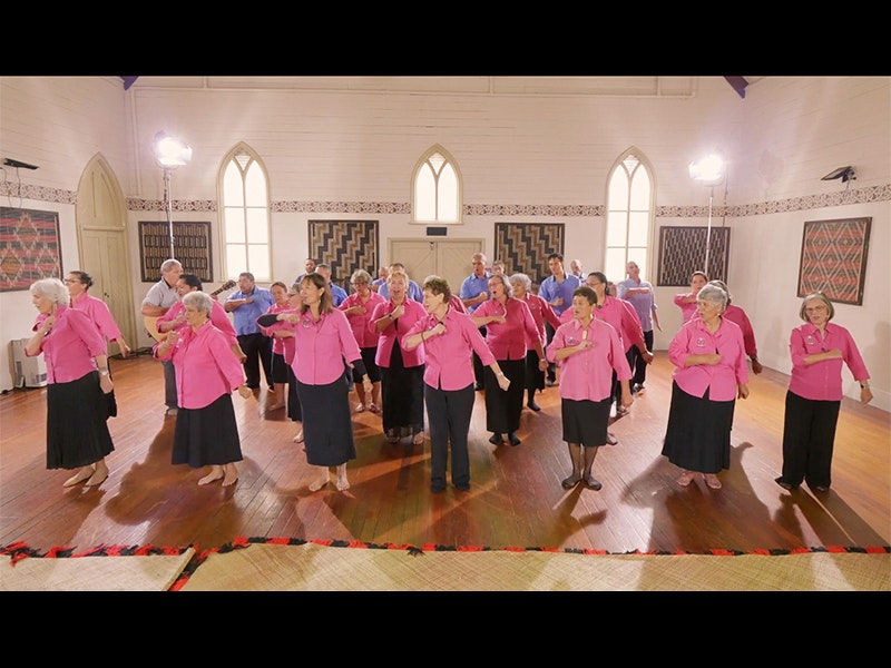 A group of older people wearing pink shirts and blue shirts dancing in formation inside a church hall