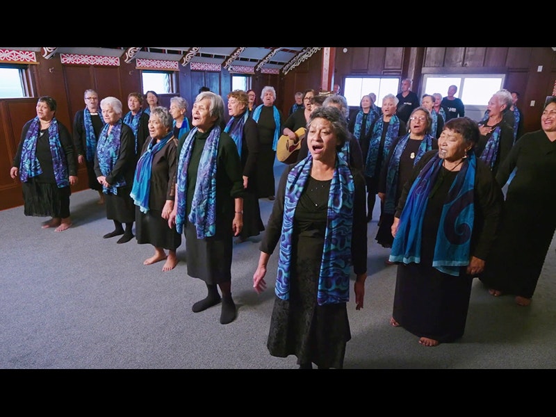 Several women in a room wearing black shirts with blue and turquoise scarves singing in unison