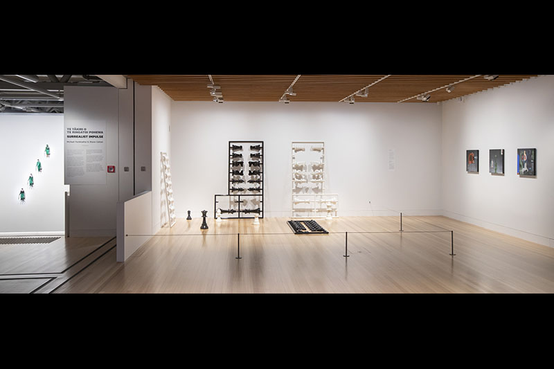 General view of the Surrealist Impulse exhibition. One work, of four men, hangs in a stairwell, one sculpture of a chess set stands on the floor leaning against the wall, and three square paintings also hang on a wall