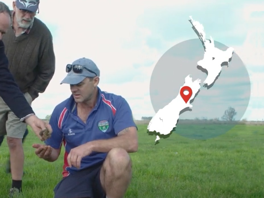 Men looking at something on the grassy ground. There's a map of New Zealand overlaid on the image on the top right-hand side.