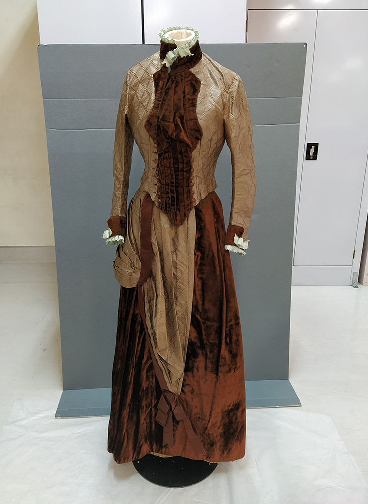 A brown velvet and silk skirt and bodice on a stand with grey cardboard behind it.