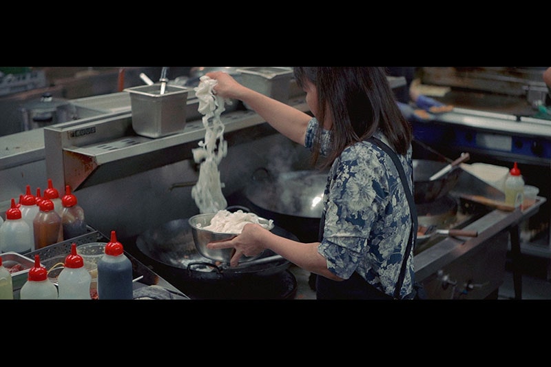 A woman is seen from behind putting noodles into a wok in a busy kitchen