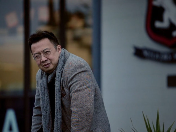 Still from the video, of Jack at St Mark’s School, Wellington, 2021. Photo by Tim Hamilton. Te Papa Jack stand with his hands on a fence that is out of shot, staring at the camera. Behind him is the crest of St Mark’s School