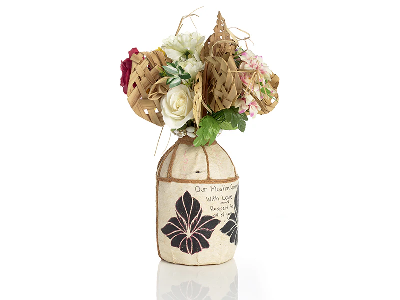 A vase of flowers and woven flax flowers with a tribute written on the side of the vase