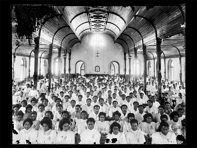 A black and white photo of a lot of Sāmoan people dressed in white and sitting in a church