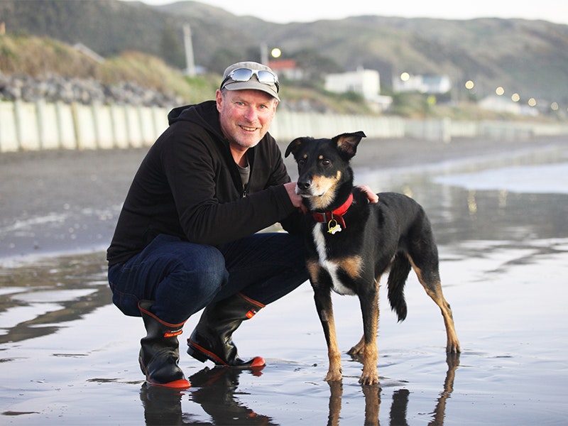 A man crouching on a beach with his hand on a dog