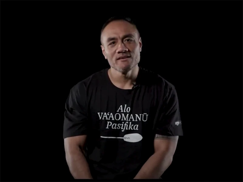 A man in a black t-shirt with Samoan writing on it sitting in a black room