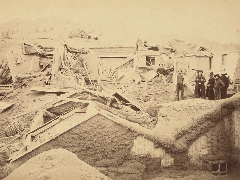 McRae’s Hotel, interior, 1886, Rotorua, Lake, by Charles Spencer. Te Papa (O.009627) Sepia photo of two people standing on the ruins of a shed covered in mud