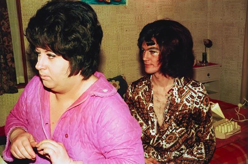 Two men, wearing wigs, sitting on a bed get dressed. One wears a pink blouse and the other a brown shirt