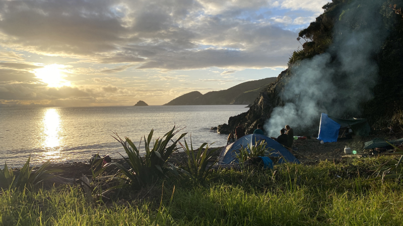 People sitting by a smoking fire on a beach at sunset.