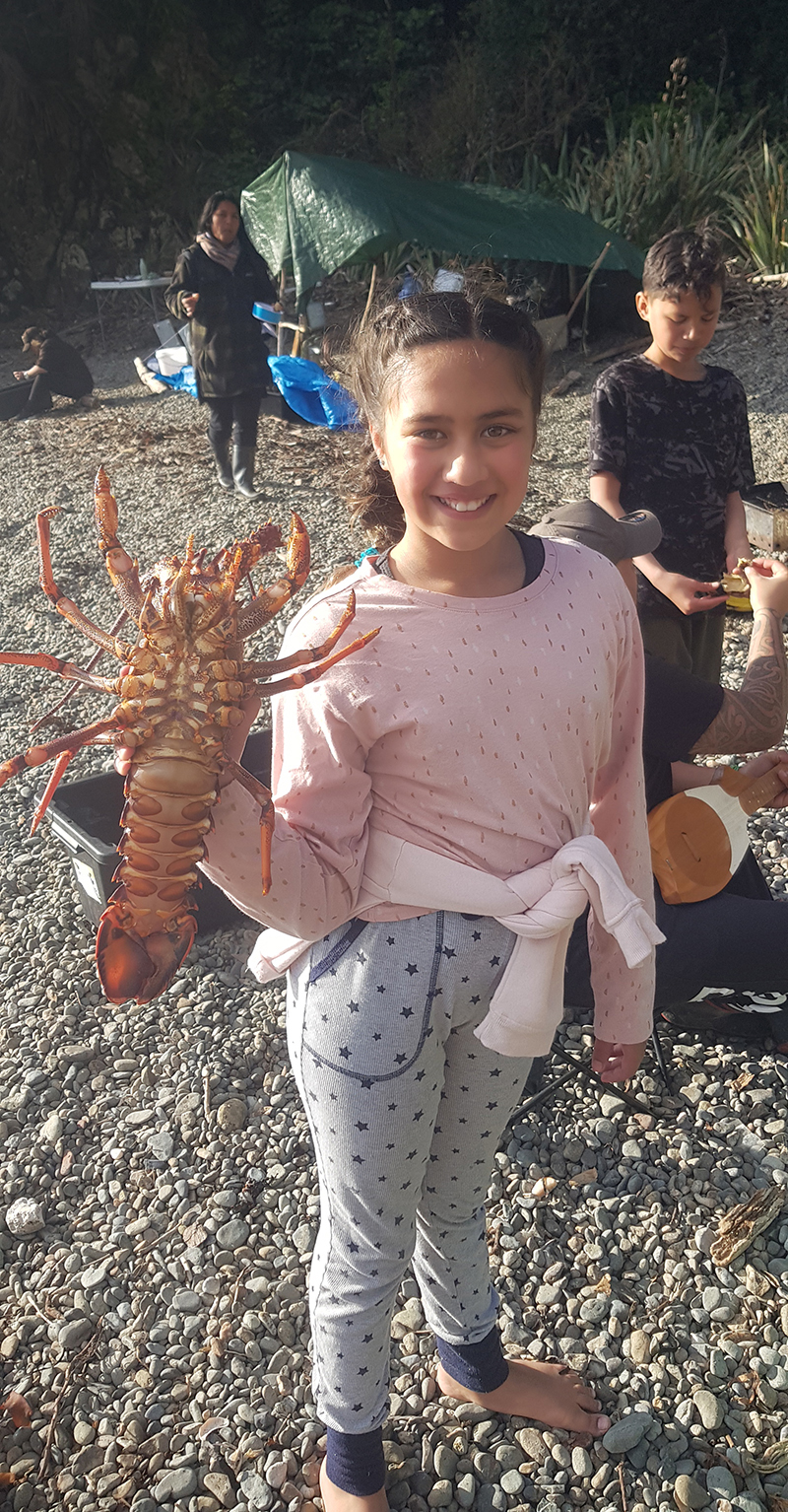 A young girl shows a large crayfish to the camera. She is standing on a rocky beach There are people behind her in the campsite.