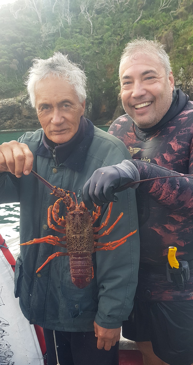 Two people standing by the ocean. One man is in a wet suit. They are holding a large crayfish and looking at the camera.