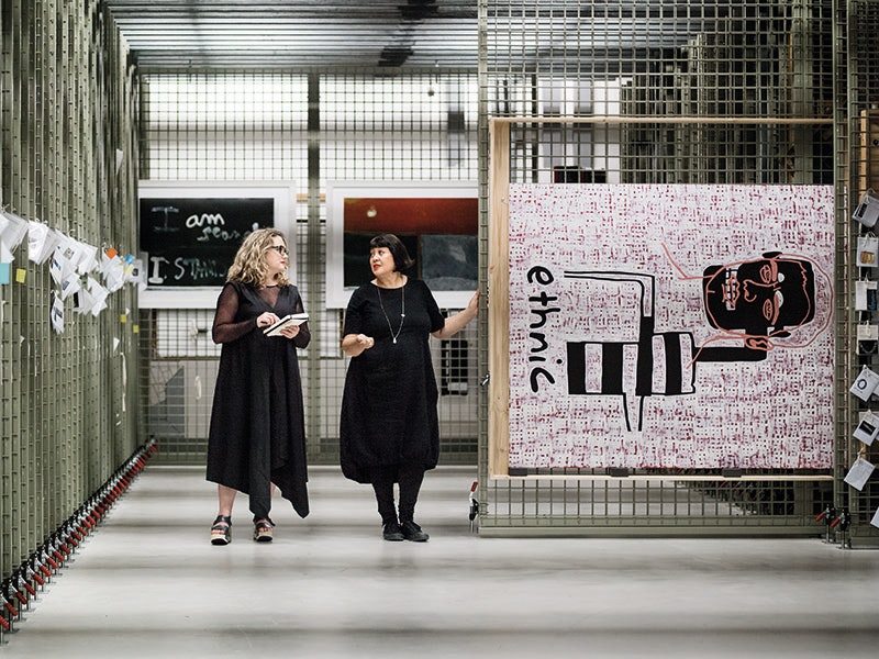 Sarah Farrar and Megan Tamati-Quennell in the paintings storeroom, with a painting on a rail pulled out and on display, with two other paintings in the background
