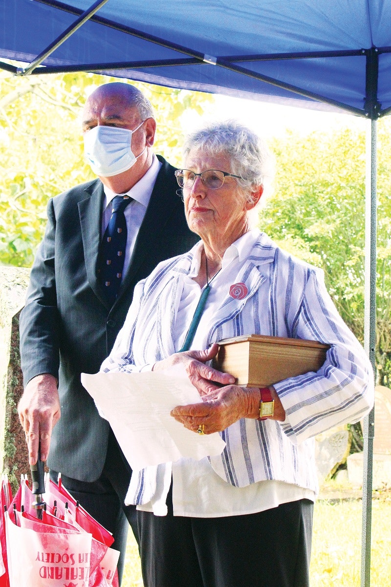An elderly man and woman stand under a marquee shelter. The woman is holding a medium-sized wooden box