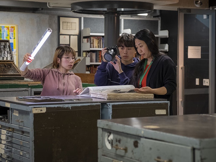 Shijia Chen (left) during a film shoot with Ya-Wen Ho (right), 2021. Photo by Daniel Crichton-Rouse. Te Papa (176043) Three people look at a large book on top of a shelving unit. One person holds a light to illuminate the book, one person is filming the scene, and another person is looking through the book