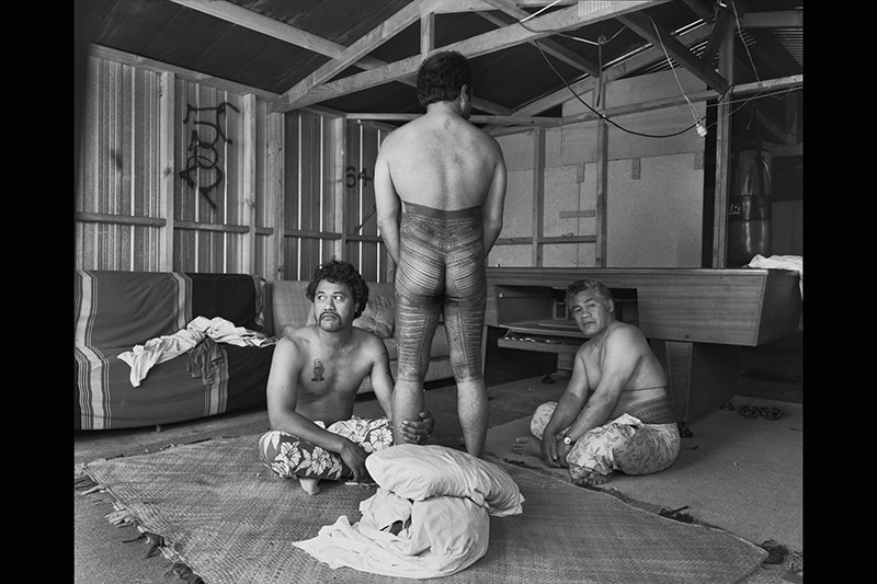 Three men with tattoos in a room. Two of the men are sitting on the floor, one is standing with his back to the camera
