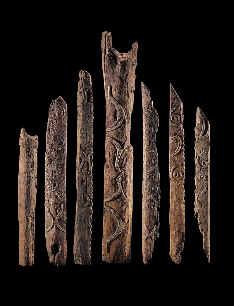 A black background with seven posts of varying height showing carvings on each one.