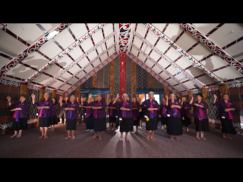 People in a group wearing the same clothing with pink and purple scarves facing the camera and singing with hand actions
