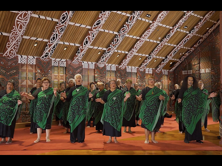 ngati_awa_tile.png A group of people in the same clothing are performing a song with actions in a carved building