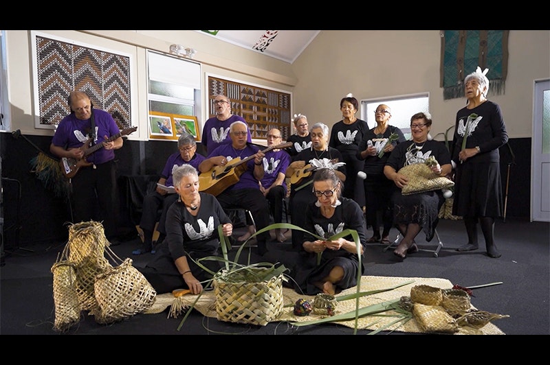Two women sitting in the foreground weaving flax with several people standing behind her singing a song. Some of those are playing a guitar and a ukulele