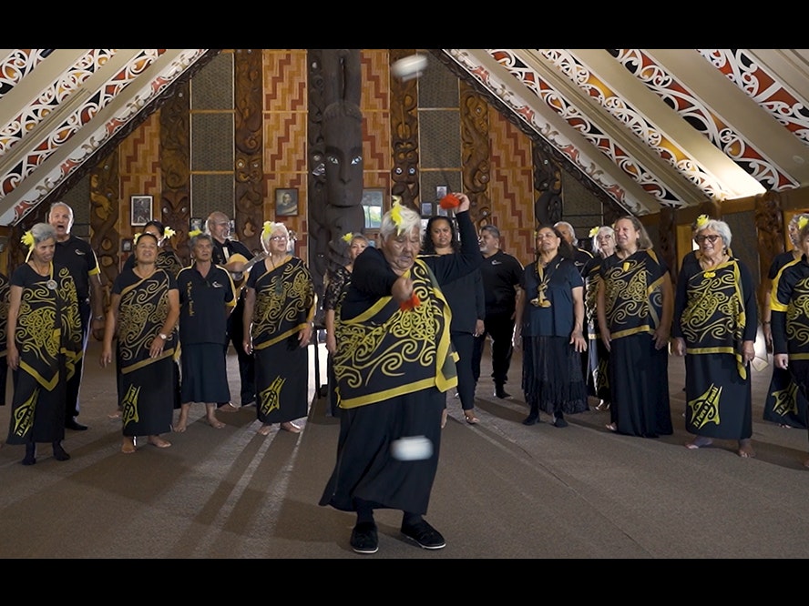 Video still from Taikura Kapa Haka 2022. Te Papa A group of people singing behind one woman doing an action song with poi (white balls on strings)