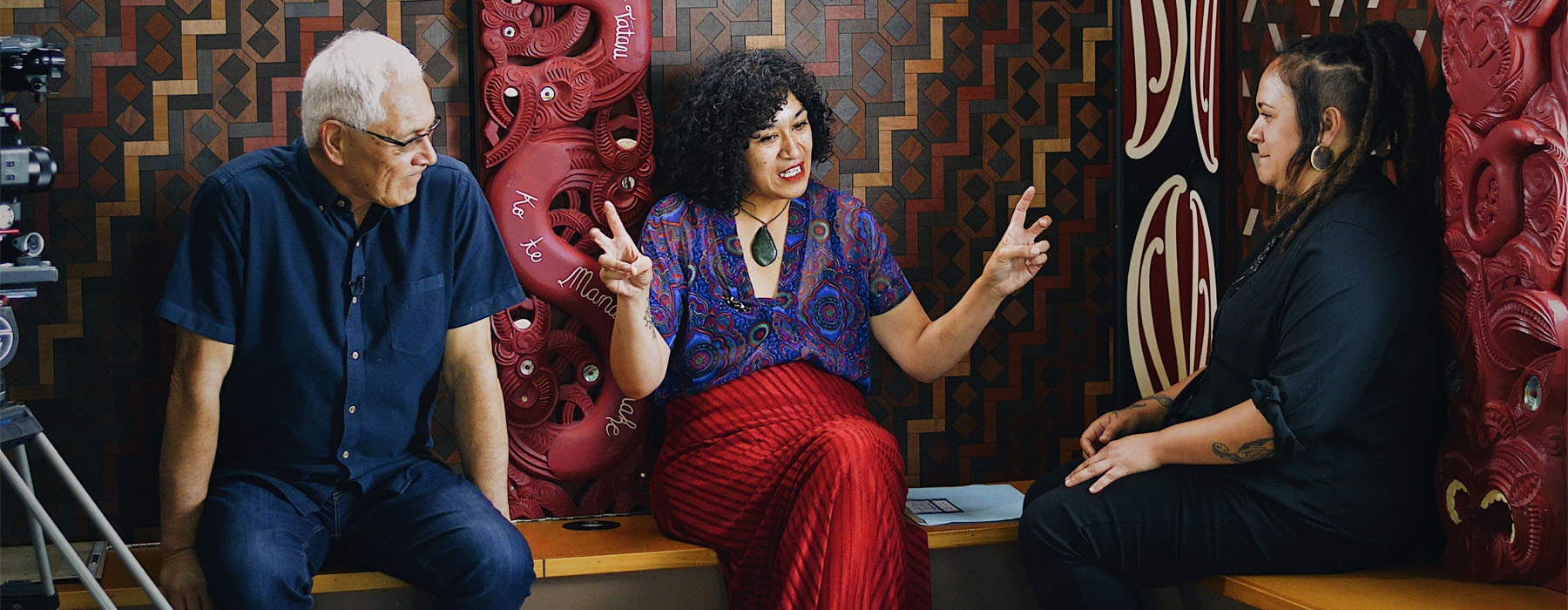Three people sit down in a marae to talk. A camera filming them can be seen partially off-screen