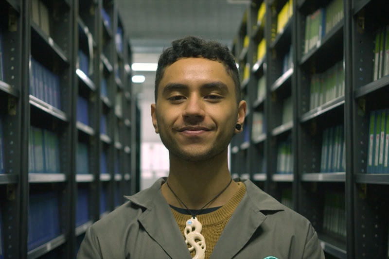 A man stands looking directly into the camera in the Ngā Taonga archives, flanked by shelving
