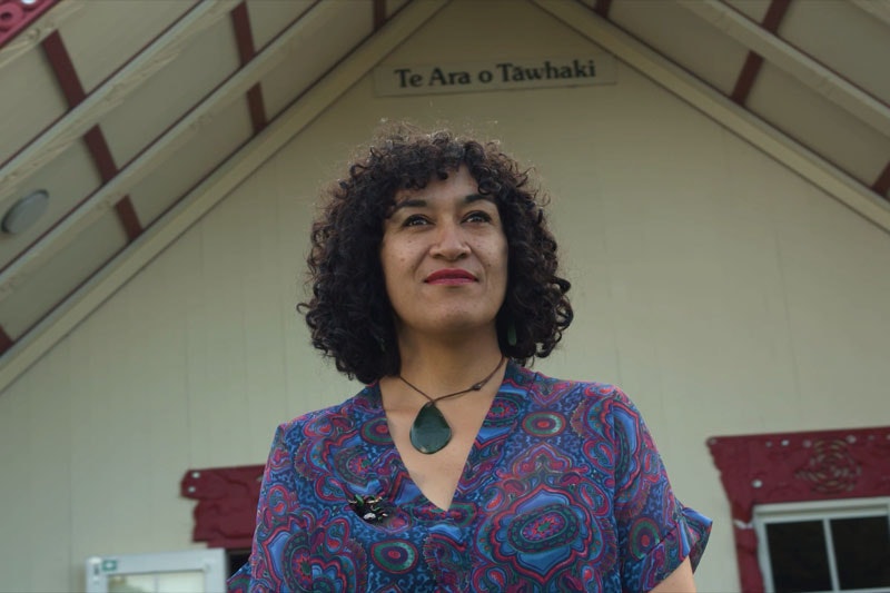 A woman stands in front of a marae looking into the camera