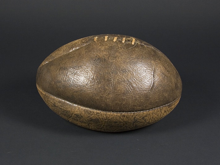 Rugby ball; about 1921; Unknown; leather ball with ink signatures. Purchased 2009. Te Papa (GH012621) A brown leather rugby ball on a black background. The ball is slightly deflated.