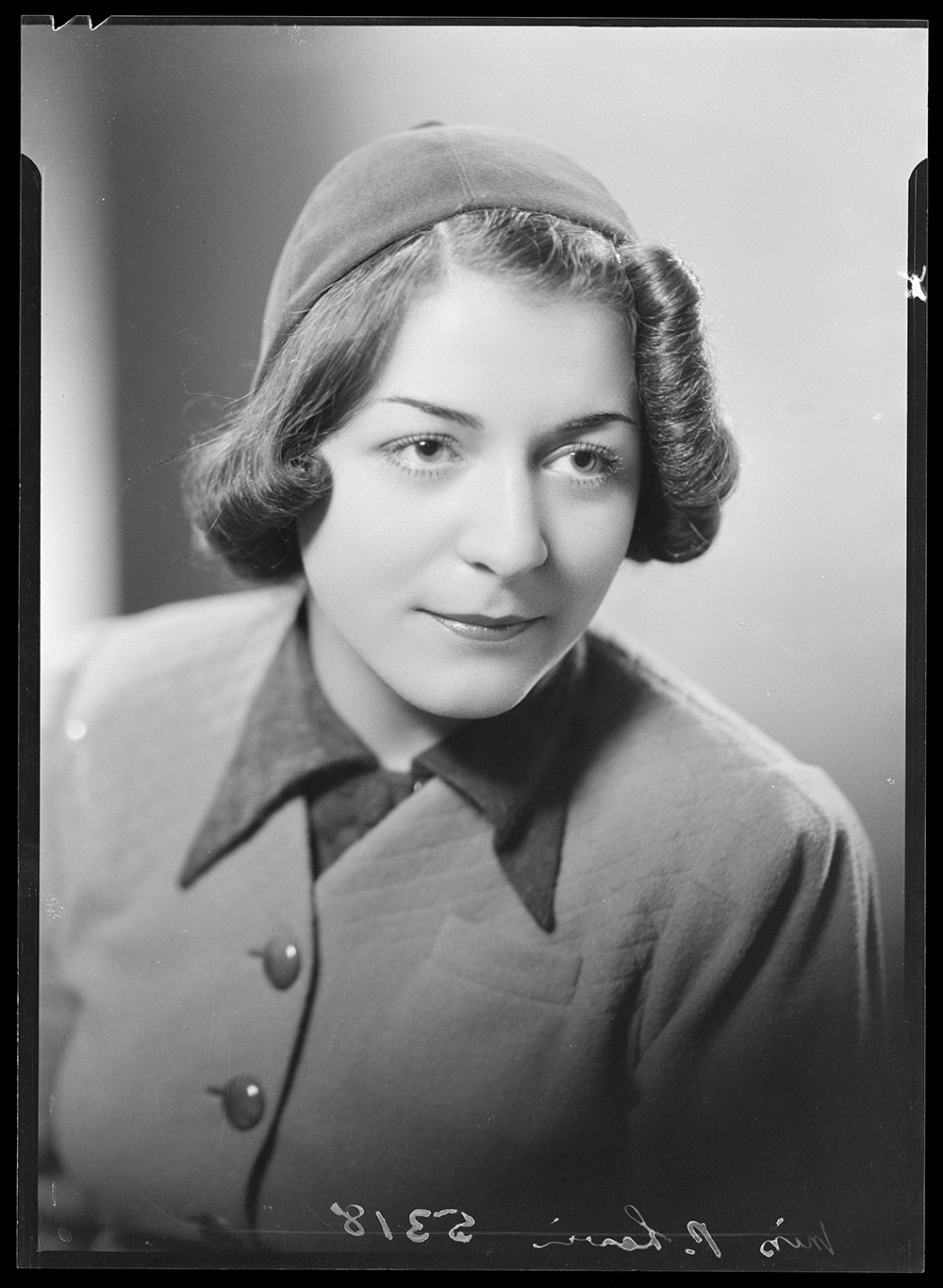 A black and white studio photograph of a woman with curled hair and wearing a hat that matches her jacket.