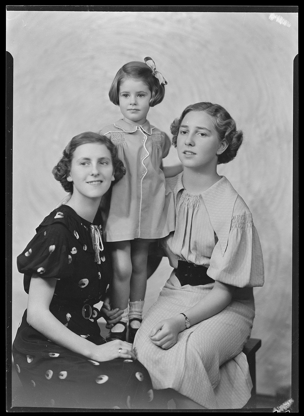Black and white photo of two women with a girl standing between them on a pedestal. They are in a photographic studio.