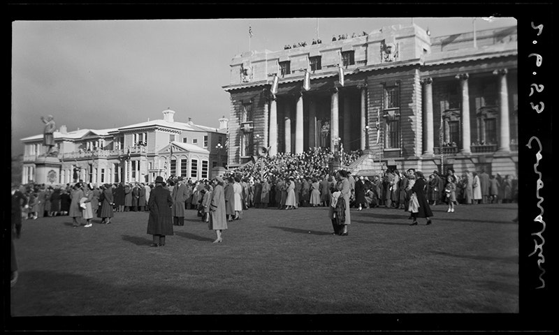 A large crowd gathering on a grassy lawn. There are a lot of people standing on the steps of a grand building