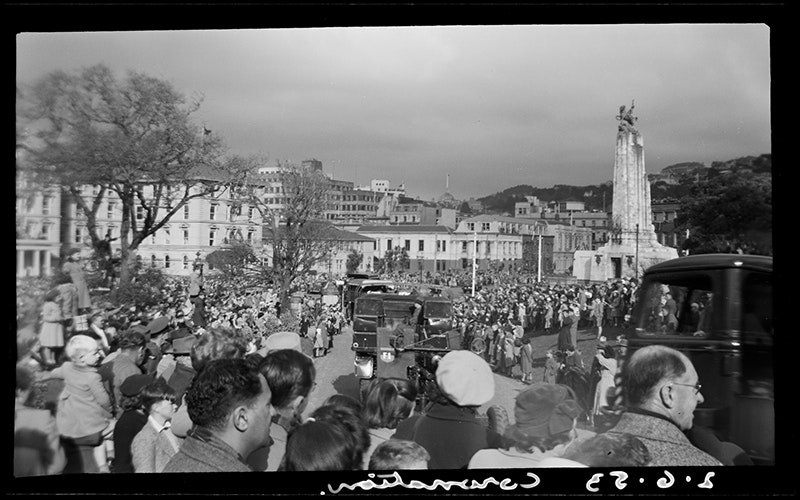 A crowd of people stand and watch a parade of military trucks. There is a tall monument and grand buildings in the background