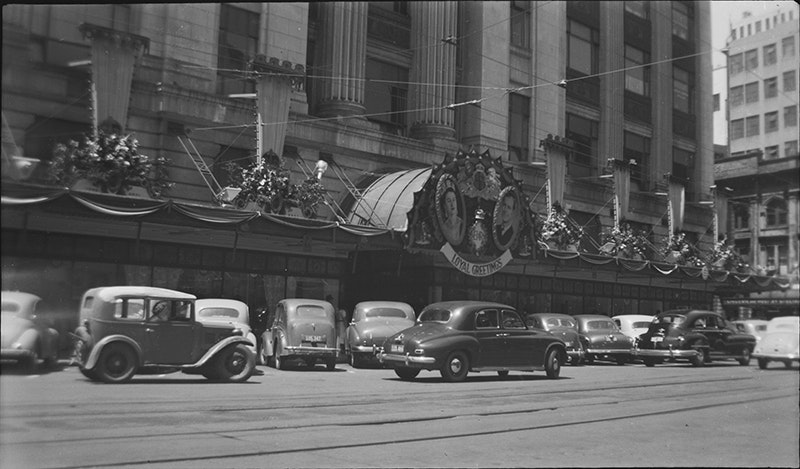Street view with 1950s cars and decorations of the Queen and Duke of Edinbugh on a building